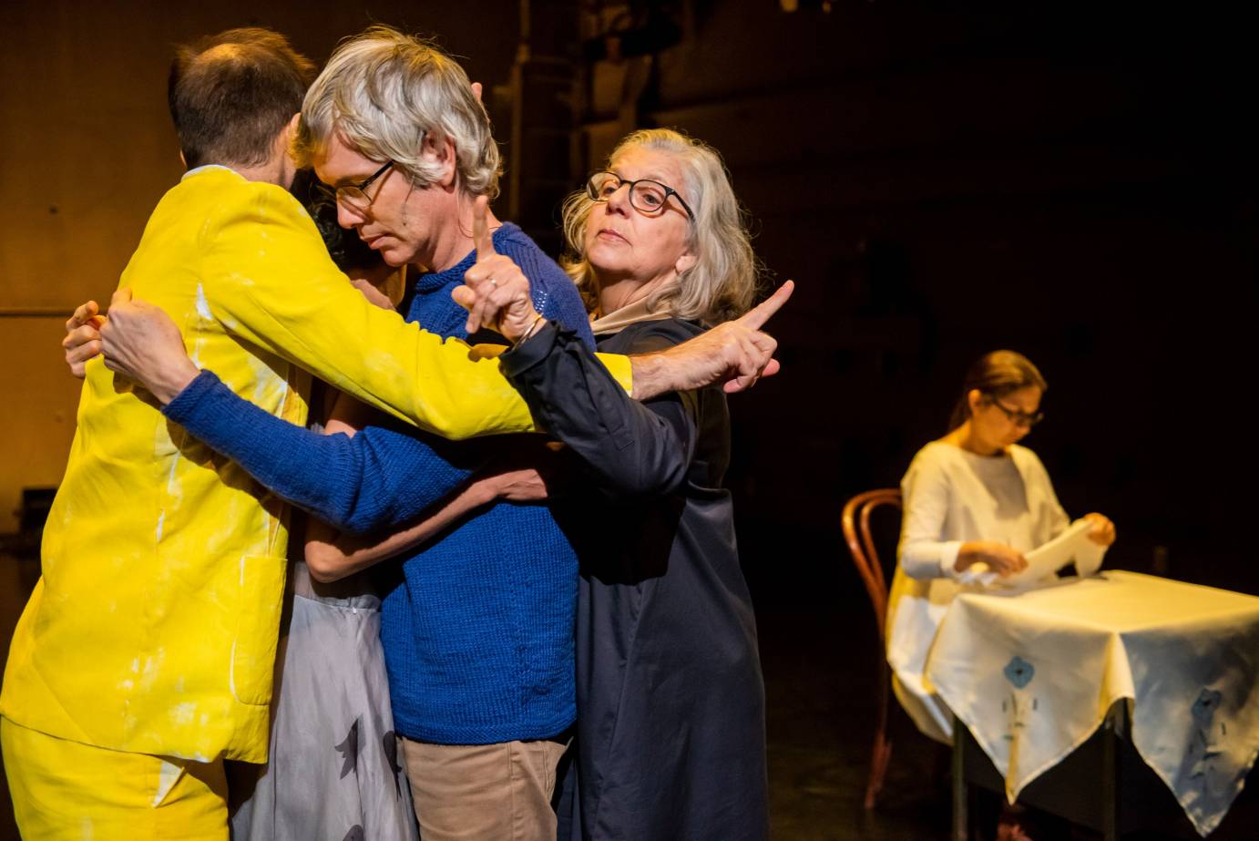 Four performers hug each other as another sits at a table folding a napkin